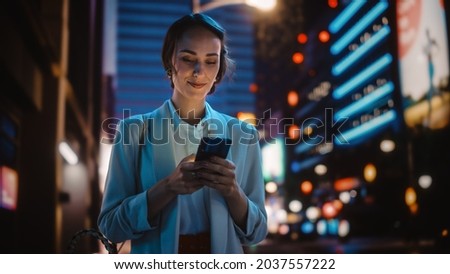 Beautiful Young Woman Using Smartphone Walking Through Night City Street Full of Neon Light. Smiling Thoughtfully Female Using Mobile Phone, Posting Social Media, Online Shopping, Texting. Royalty-Free Stock Photo #2037557222