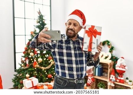 Senior man with beard wearing santa claus hat celebrating christmas at home, standing by christmas tree holding present taking a selfie picture