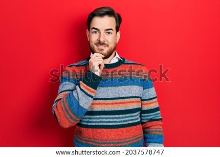 Handsome caucasian man with beard wearing elegant wool winter sweater looking confident at the camera with smile with crossed arms and hand raised on chin. thinking positive. 