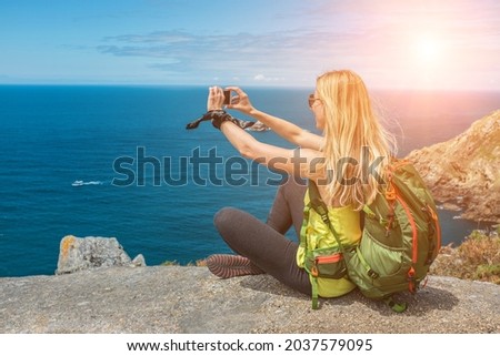 Woman hiker, hiking backpacker traveler camper with her phone camera on the top of mountain in sunny day under sun light. Beautiful sea landscape view.