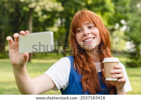 Smiling caucasian student young woman girl taking selfie photo on smart phone, having video call with friends, posting on social media, vlogging and blogging online in park drinking coffee.
