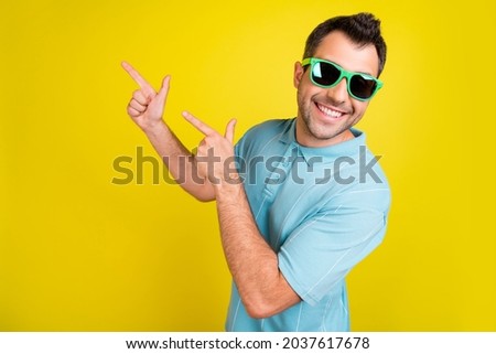 Photo of funky pretty young guy dressed blue t-shirt dark glasses smiling pointing empty space isolated yellow color background