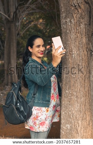A vertical shot of a young Latin female taking a selfie with a phone next to the tree