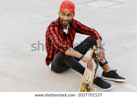 Smiling young african man in casual wear sitting with skateboard outside on skate ramp