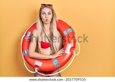 Young caucasian woman wearing bikini and holding lifeguard float making fish face with lips, crazy and comical gesture. funny expression. 