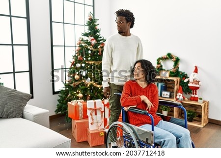 Young interracial couple with woman sitting on wheelchair by christmas tree looking to side, relax profile pose with natural face with confident smile. 