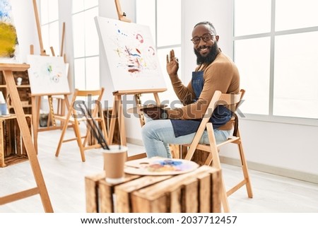 African american artist man painting on canvas at art studio doing ok sign with fingers, smiling friendly gesturing excellent symbol 