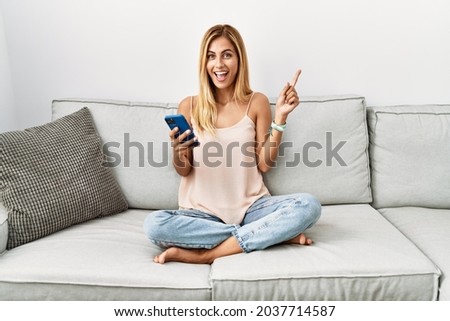 Blonde beautiful young woman sitting on the sofa at home using smartphone with a big smile on face, pointing with hand finger to the side looking at the camera. 