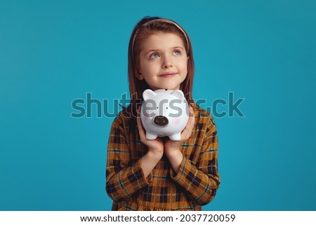 Cute adorable girl kid holding money box in shape of pig near face looking up and dreaming about something, isolated over blue studio background.