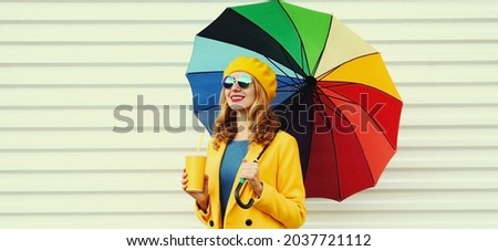 Autumn portrait of happy cheerful smiling young woman with colorful umbrella and cup of juice wearing a yellow coat and beret on a white background