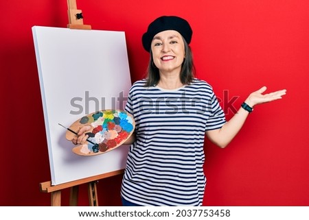 Middle age hispanic woman standing drawing with palette by painter easel stand smiling cheerful presenting and pointing with palm of hand looking at the camera. 