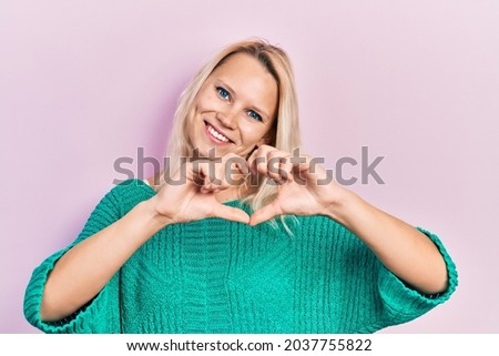 Beautiful caucasian blonde woman wearing casual winter sweater smiling in love doing heart symbol shape with hands. romantic concept. 