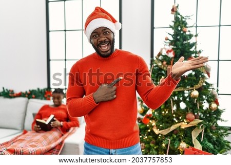 Young african american man standing by christmas tree amazed and smiling to the camera while presenting with hand and pointing with finger. 