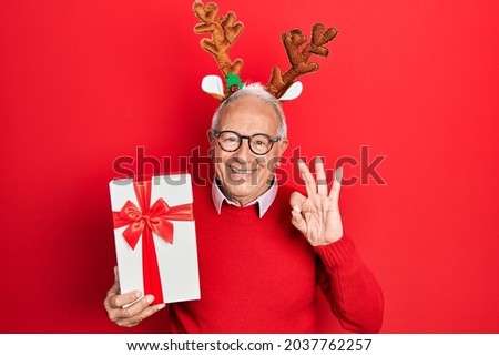 Senior man with grey hair wearing deer christmas hat holding gift doing ok sign with fingers, smiling friendly gesturing excellent symbol 