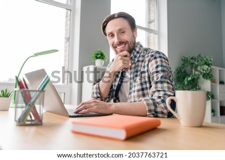 Photo of adorable thoughtful young man wear plaid shirt smiling sitting table chatting modern gadget indoors house office