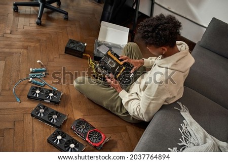 Top view of African American man sit on floor check hardware to upgrade his farm. Millennial ethnic male install video card adapter to motherboard. Technology, mining, farming concept. 