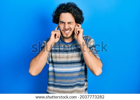 Handsome hispanic man wearing casual clothes over pink background covering ears with fingers with annoyed expression for the noise of loud music. deaf concept. 