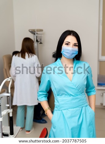 Woman doctor wearing protective mask stands with arms in pockets and looks at camera. Modern clinic with up-to-date equipment for eye testing. Closeup