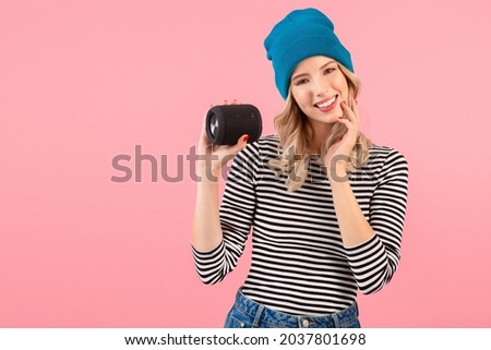 young pretty woman holding wireless speaker listening to music wearing striped shirt and blue hat smiling happy positive mood posing on pink background isolated