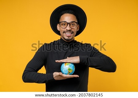 Young happy geographer excited fun african man 20s wear stylish black shirt hat eyeglasses holding in palms Earth world globe looking camera isolated on yellow orange color background studio portrait.