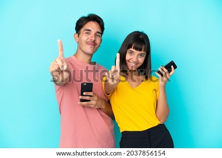Young mixed race couple holding mobile phone isolated on blue background showing and lifting a finger