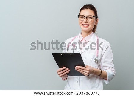 Portrait of a friendly smiling young female doctor. Image of a female doctor with glasses and with a document folder. Isolated on a gray background. Royalty-Free Stock Photo #2037860777