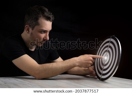 A young handsome man in black clothes points his finger at the center of a glowing target on a black background in a low key. Goal achievement concept