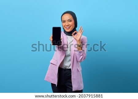 Cheerful Asian woman showing smartphone blank screen and gesturing okay sign over blue background