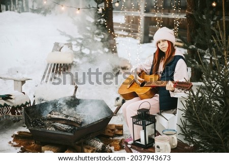 teenage girl in a vest and a knitted hat plays the guitar outdoor near a fire pit, the concept of winter Christmas holidays and active lifestyle