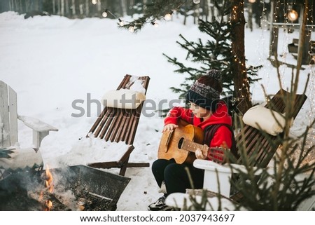 boy in a vest and a knitted hat plays the guitar outdoor near a fire pit, the concept of winter Christmas holidays and active lifestyle
