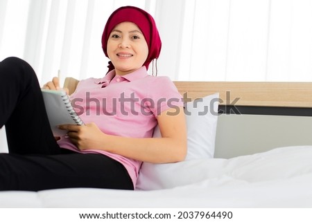 Asian woman on pink shirt, black trousers, cover head by red scarf as breast cancer patient, up knee to take sad note on paper while relaxing on house bed beside alarm clock, photo frame, plant pot
