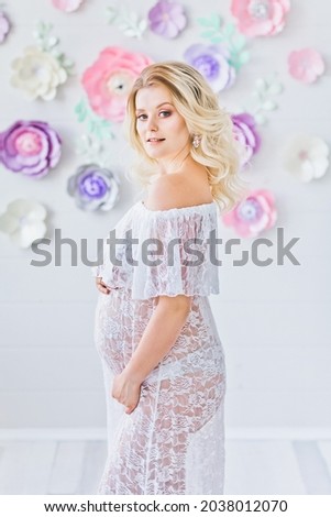 Smiling pregnant woman posing near white wall with decorative flowers. Happy pregnant blonde in white interior.  Happy motherhood.