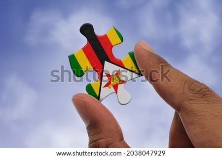 Hand holding piece of jigsaw puzzle with flag of Zimbabwe. Jigsaw puzzle of Zimbabwe flag on sky background.