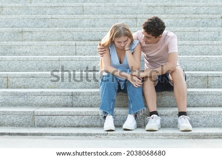 Teenager couple sitting on the stairs. Hispanic boyfriend trying to cheer up and hugging sad girlfriend. Relationship troubles, anxiety and depression in adolescence concept with copy space.