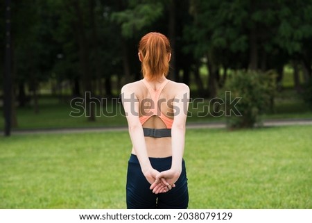Attractive redhead young woman in sportswear stretching her shoulders in summer in green park. Back view. The girl is warming up. Concept of sport, healthy lifestyle and exercise. Royalty-Free Stock Photo #2038079129