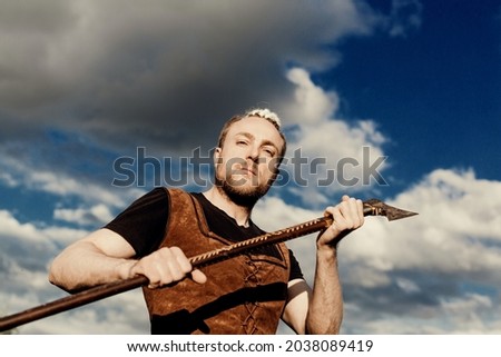 A young man with an interesting depicts a Viking with an ax in an unreliable stylized modern costume against the background of the sky and clouds