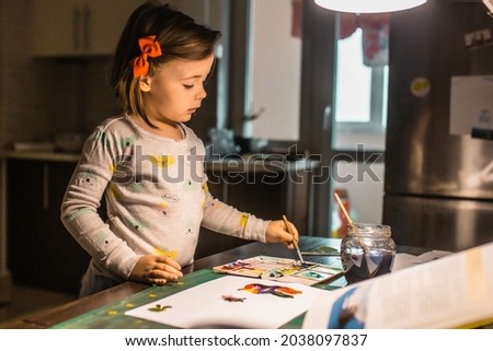 Cute little girl learns how to paint at the table. Adorable preschooler girl painting with watercolor in the evening. Art classes concept. Preschooler education theme.