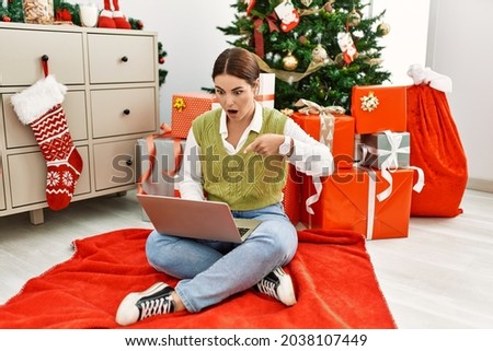 Young beautiful hispanic woman using laptop sitting by christmas tree pointing down with fingers showing advertisement, surprised face and open mouth 
