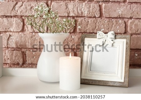 Burning candle with picture frame and vase on table near brick wall