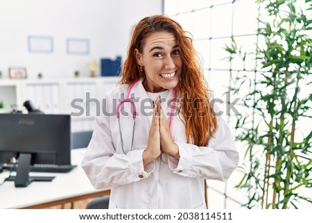 Young redhead woman wearing doctor uniform and stethoscope at the clinic praying with hands together asking for forgiveness smiling confident. 