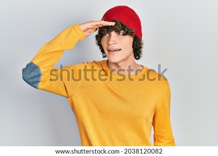 Handsome young man wearing wool hat very happy and smiling looking far away with hand over head. searching concept. 