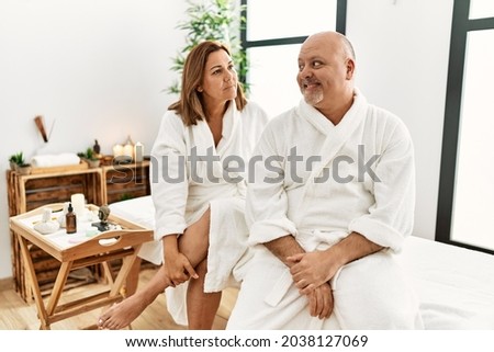 Middle age hispanic couple wearing bathrobe at wellness spa looking to side, relax profile pose with natural face with confident smile. 