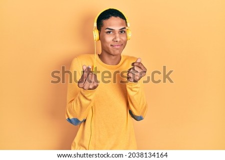 Young african american guy listening to music using headphones doing money gesture with hands, asking for salary payment, millionaire business 