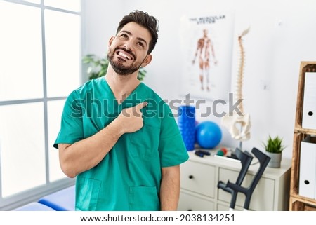 Young man with beard working at pain recovery clinic cheerful with a smile of face pointing with hand and finger up to the side with happy and natural expression on face 