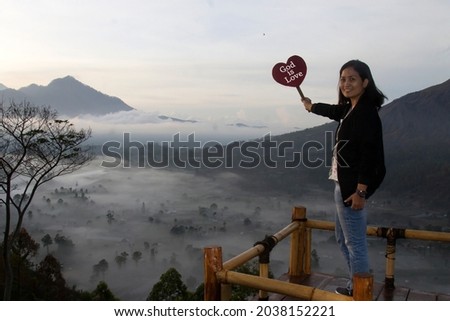 God is Love. Happy woman standing with a smile holding red love heart sign in the mountains at sunrise on a misty morning. Love and happiness concepts.