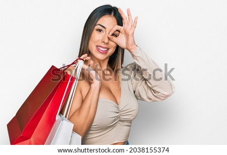 Young hispanic woman holding shopping bags smiling happy doing ok sign with hand on eye looking through fingers 