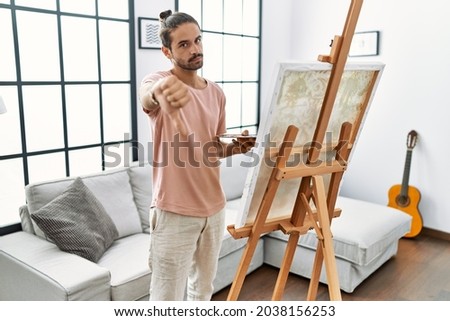 Young hispanic man with beard painting on canvas at home looking unhappy and angry showing rejection and negative with thumbs down gesture. bad expression. 
