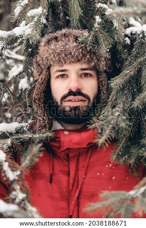 Portrait of a bearded man dressed in a warm winter hat-earflaps standing in the middle of the forest of snow-covered trees