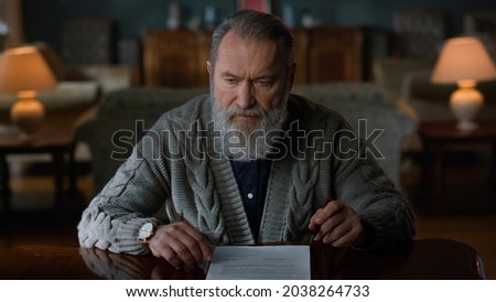 Pensive senior man doing paperwork in luxury living room. Portrait old business man signing document in vintage cabinet. Elderly businessman working with papers in home interior.