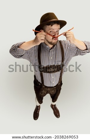 Cute bearded man, waiter in traditional Austrian or Bavarian costume holding smoked sausages isolated over gray background. Funny meme emotions. Celebration, oktoberfest, festival concept.